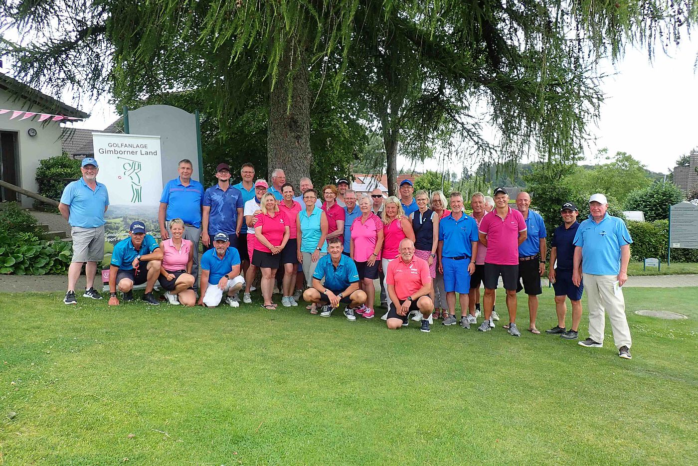 Männer und Frauen nach dem Golf Spielen in pink und blau als Gruppe vor einem Baum.